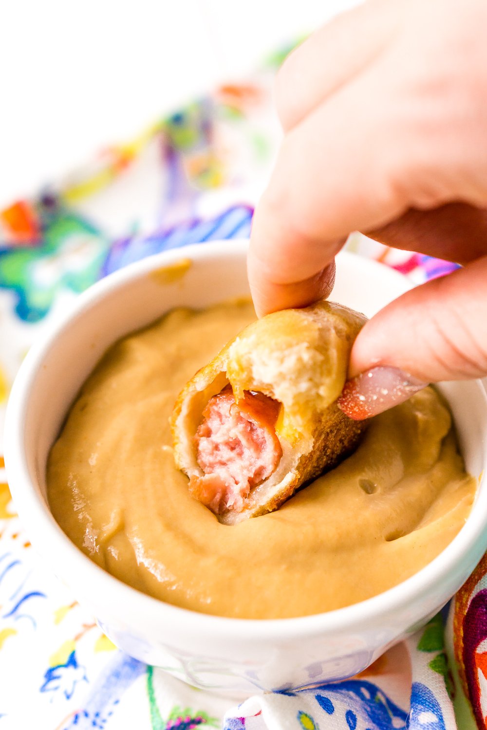 Woman's hand dipping pigs in a blanket into dipping sauce with bite taken out of it.