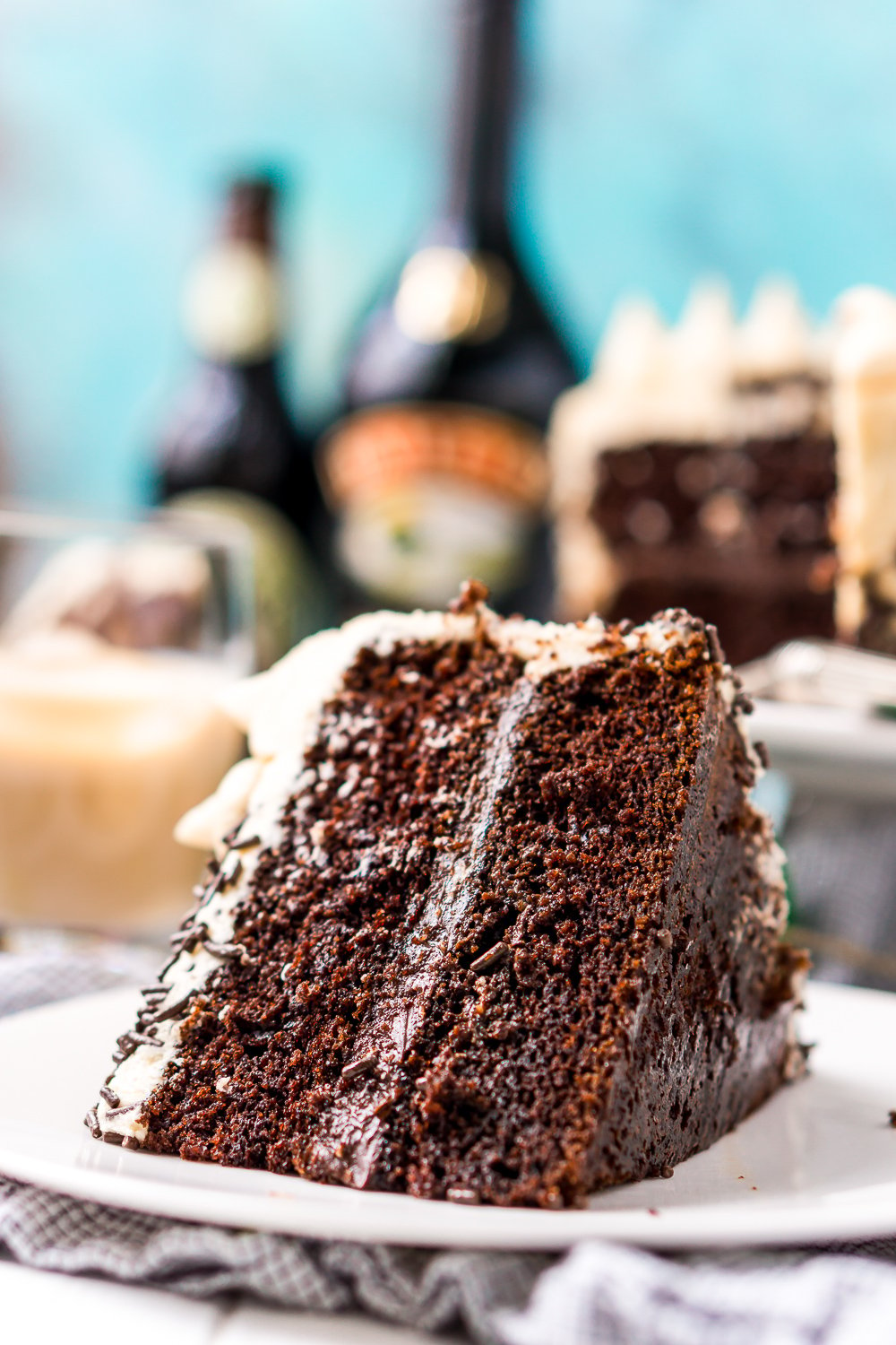 Slice of Chocolate Guinness Cake on a white plate with the rest of the cake, bottle of stout and irish cream in the background.