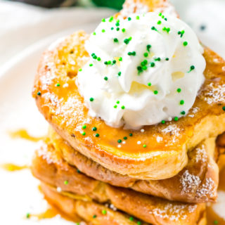 Slightly overhead photo of a stack of Irish Cream French Toast on a white plate with caramel syrup whipped cream and green sprinkles.