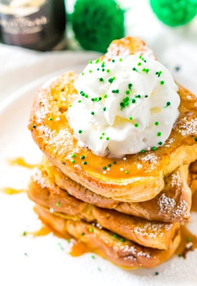 Slightly overhead photo of a stack of Irish Cream French Toast on a white plate with caramel syrup whipped cream and green sprinkles.