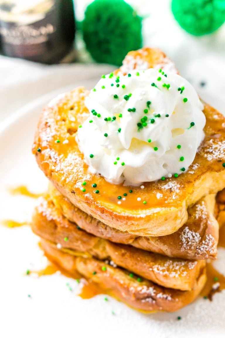 Slightly overhead photo of a stack of Irish Cream French Toast on a white plate with caramel syrup whipped cream and green sprinkles.
