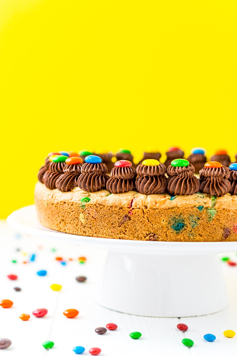 M&M's Cookie Cake with chocolate frosting on a white cake stand with yellow background.