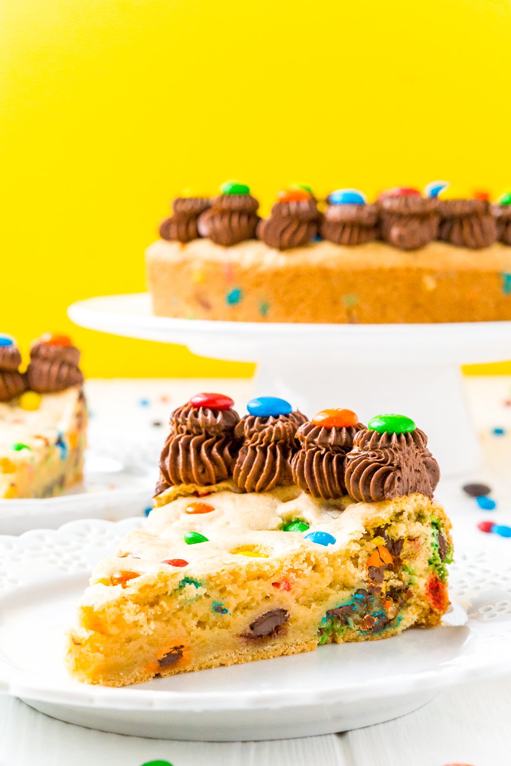 Slice of M&M's Cookie Cake on white plate with rest of cake in the background.