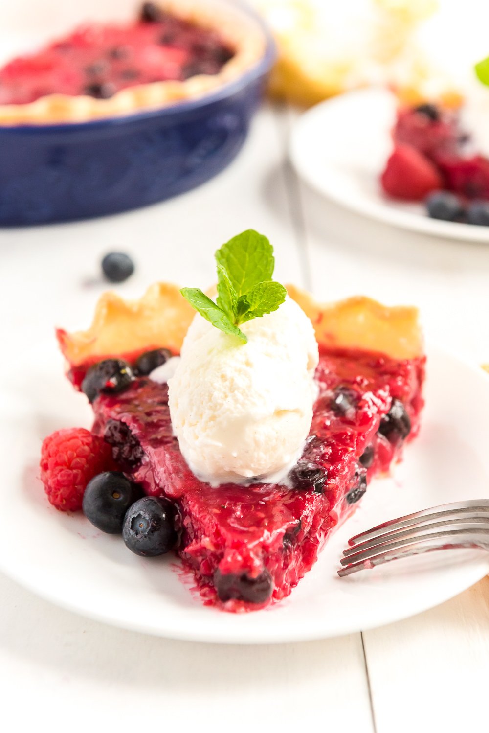 Straight on photo of a slice of Mixed Berry Pie pointed towards the camera with scoops of vanilla ice cream on top and fresh berries on the side.