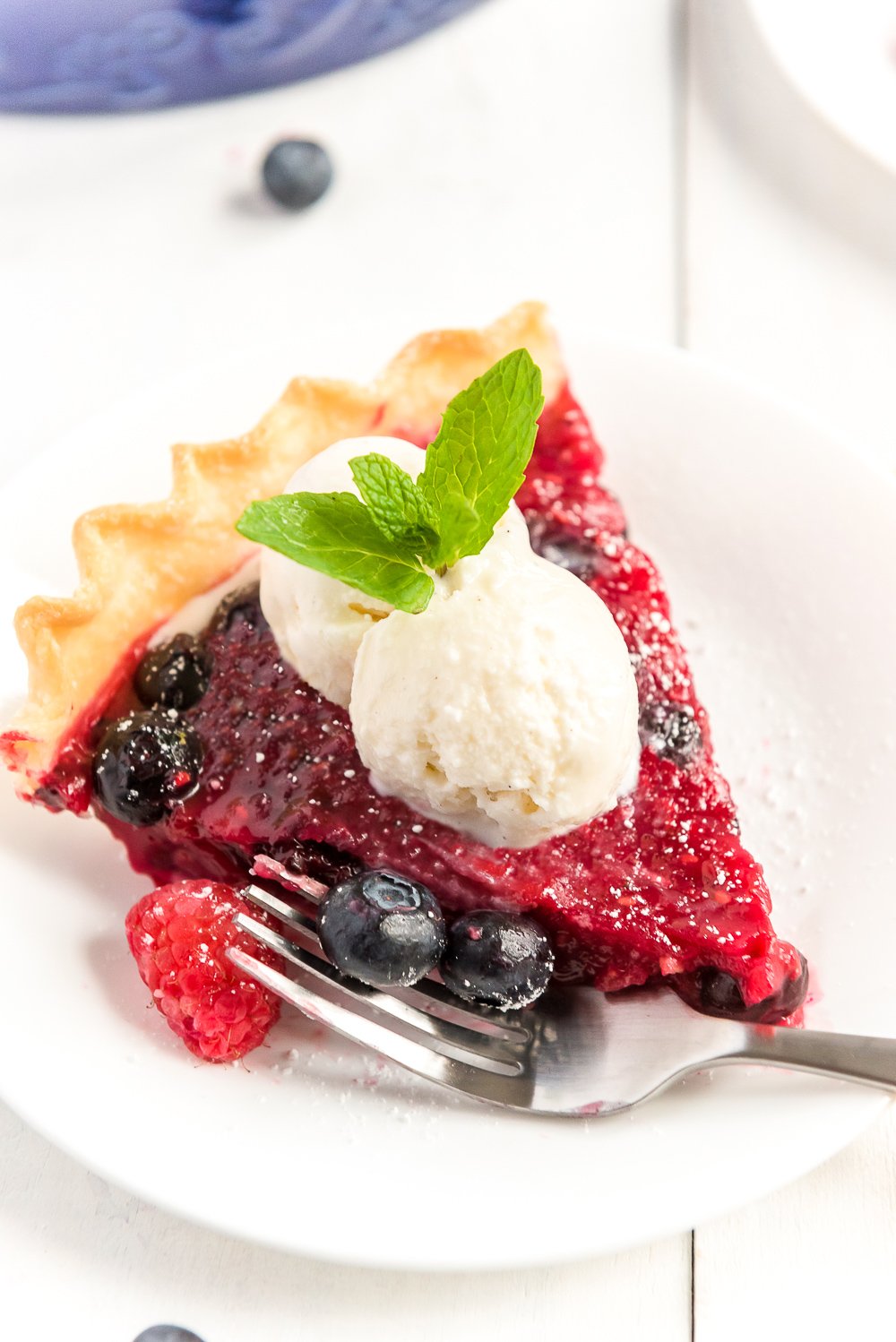 Angled photo of a slice of Mixed Berry Pie on a white plate with scoops of vanilla ice cream on top.