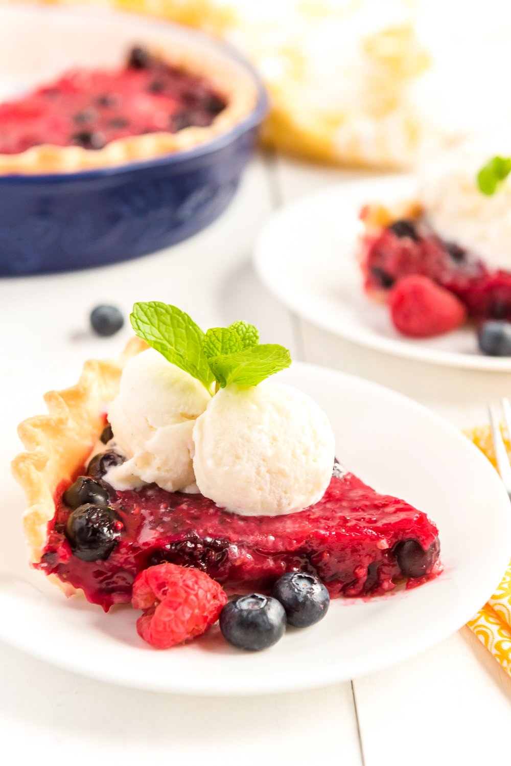 Slice of Mixed Berry Pie on a white plates with fresh berries, vanilla ice cream on top, and a sprig of mint.