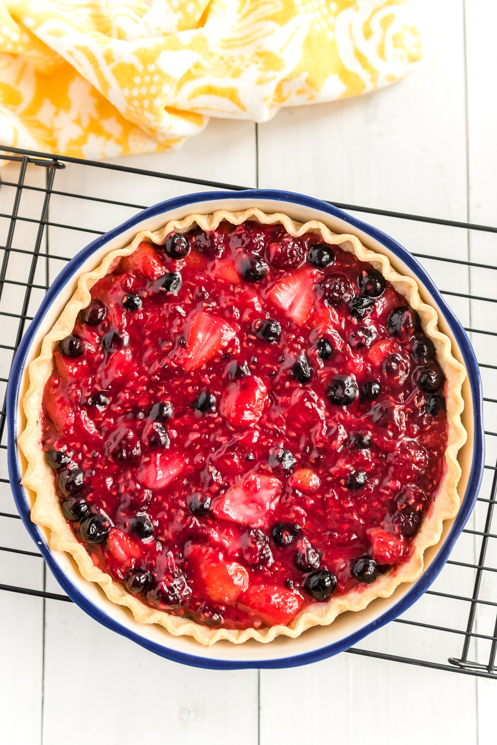 Mixed Berry Pie on black cooling rack on white wood with yellow napkin.