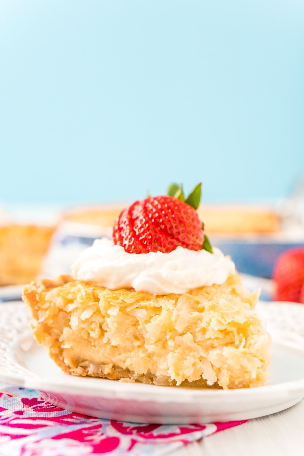 Slice of Coconut Custard Pie on a white plate with pink and white napkin and a blue background. Pie has whipped cream and a strawberry on top.