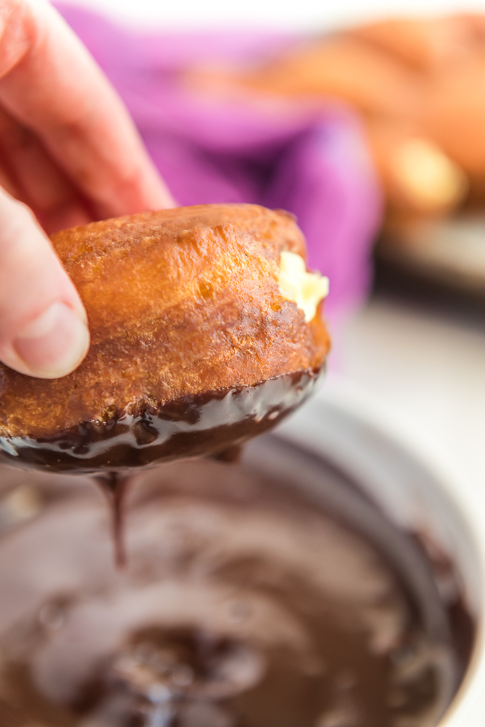 Donut filled with pastry cream being dipped in chocolate ganache.