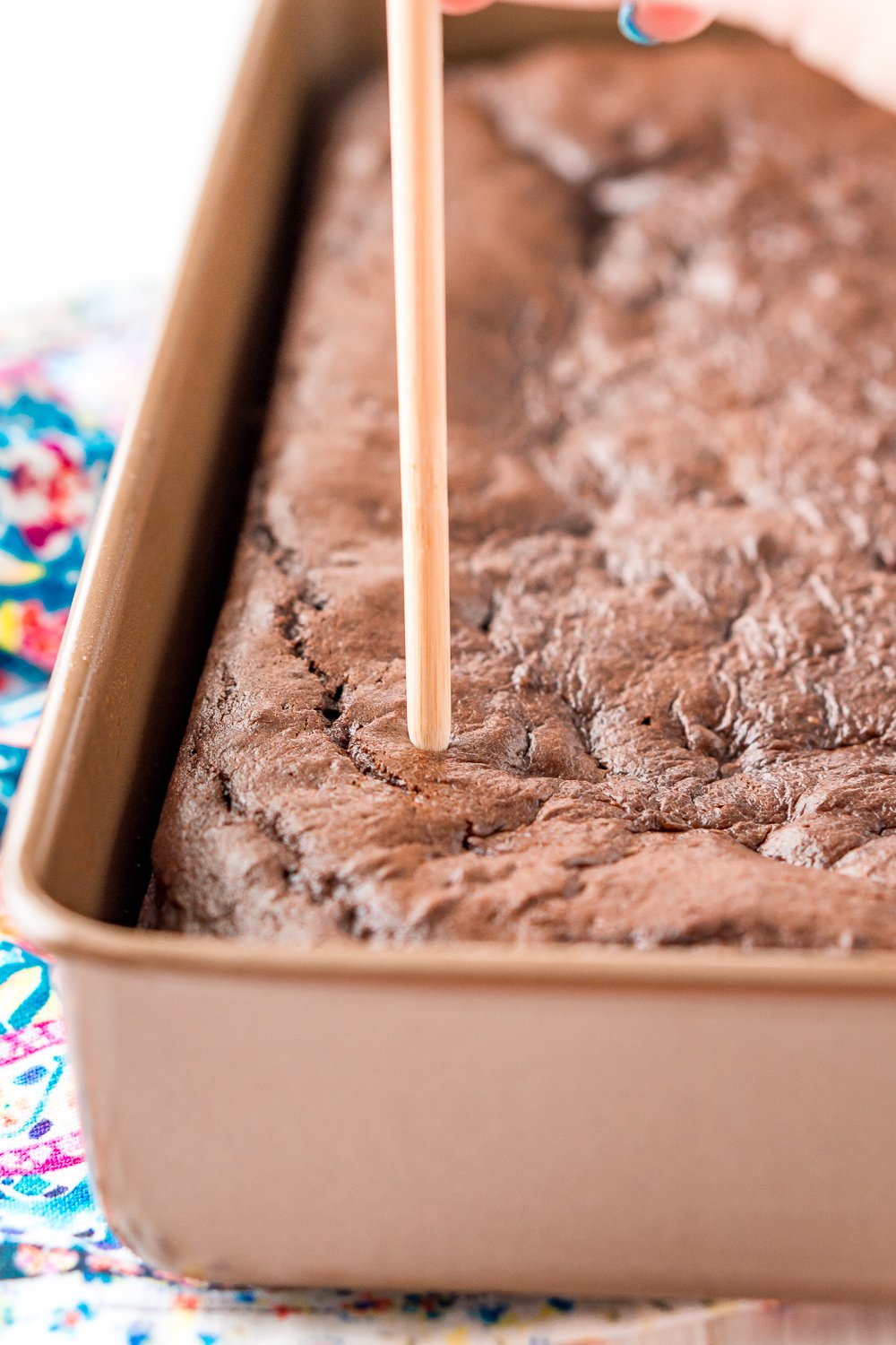 Chocolate cake in a 9x13-inch pan with a woman's hand holding a wooden skewer poking holes in the cake.
