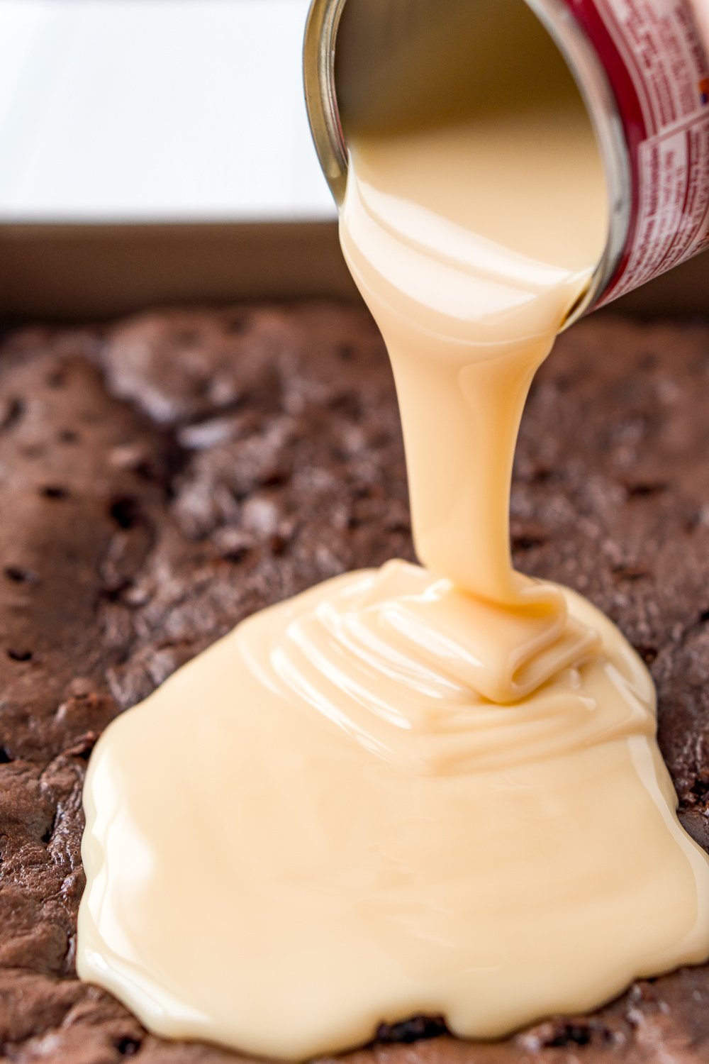 Can of sweetened condensed milk being poured over a chocolate cake.