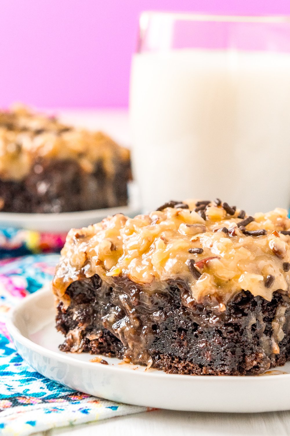 Slice of German Chocolate poke Cake on a small white plate with a glass of milk and another slice of cake in the background.