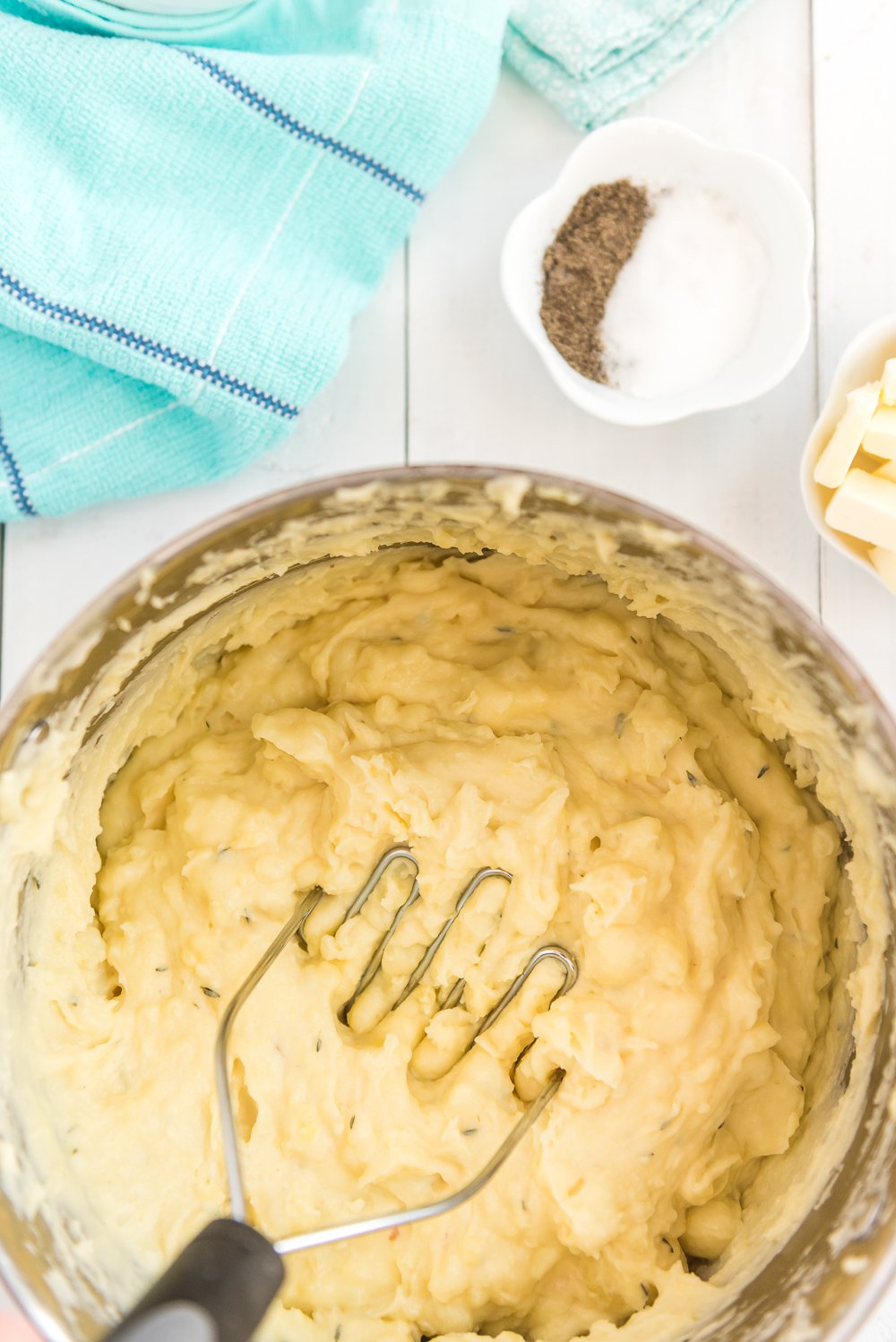 Overhead photo of boiled potatoes in large pot being mashed with a potato masher. Blue napkin and bowl of salt and pepper and butter to the side.