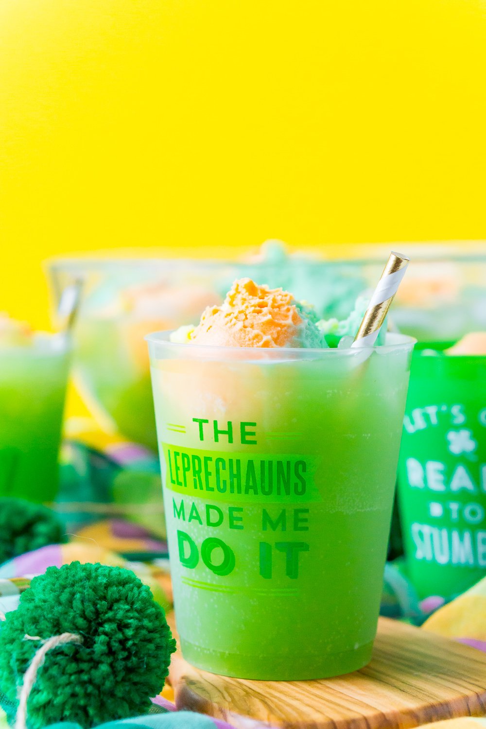 Cup of St. Patrick's Day Punch that says " The Leprechauns Made Me Do It" on a wood board with a punch bowl and more cups in the background.