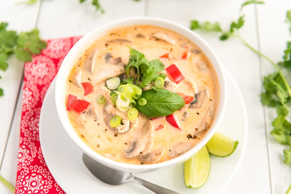 Chicken Coconut Soup in a white bowl on a white wooden table with a red napkin and lime wedges scattered around.