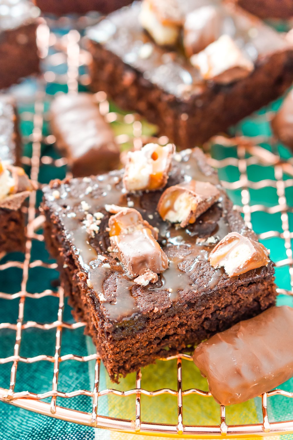 A square Twix Caramel Brownie on a copper cooling rack with more brownies and mini Twix bars surrounding it.