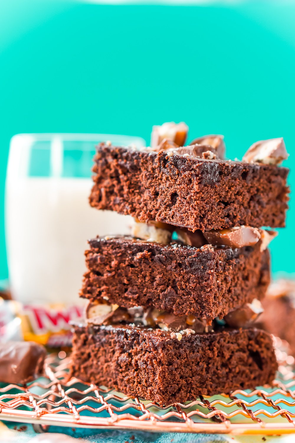 Stack of three Twix Caramel Brownies on a copper cooling rack with a glass of milk in the background.