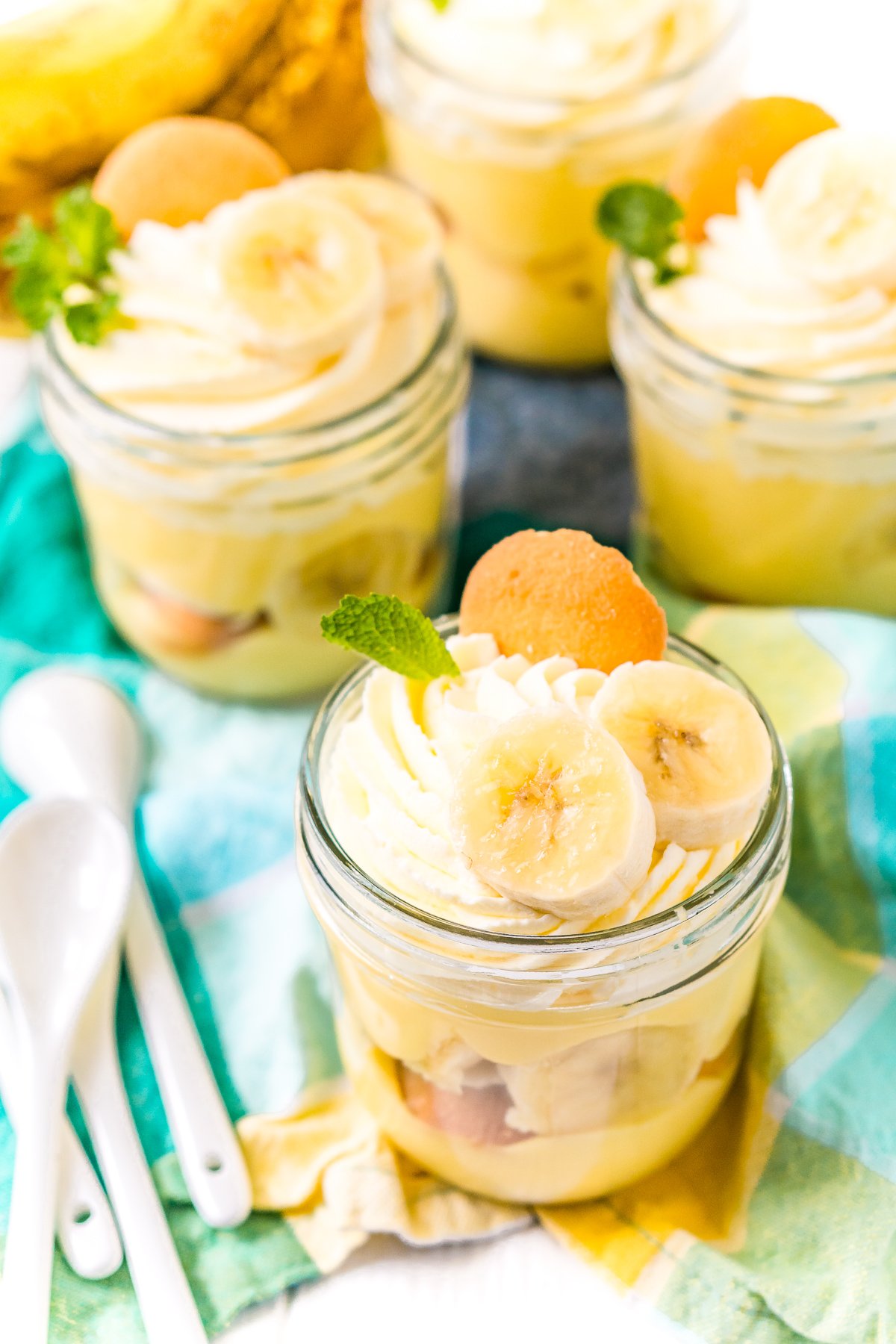 Mini mason jars with banana pudding in them on a blue and yellow napkin with white spoons to the side.