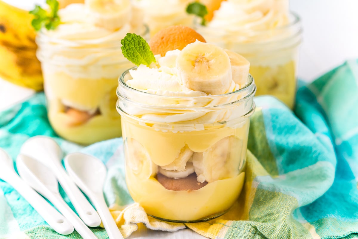 Three mason jars filled with banana pudding on a blue napkin with white spoons to the side.