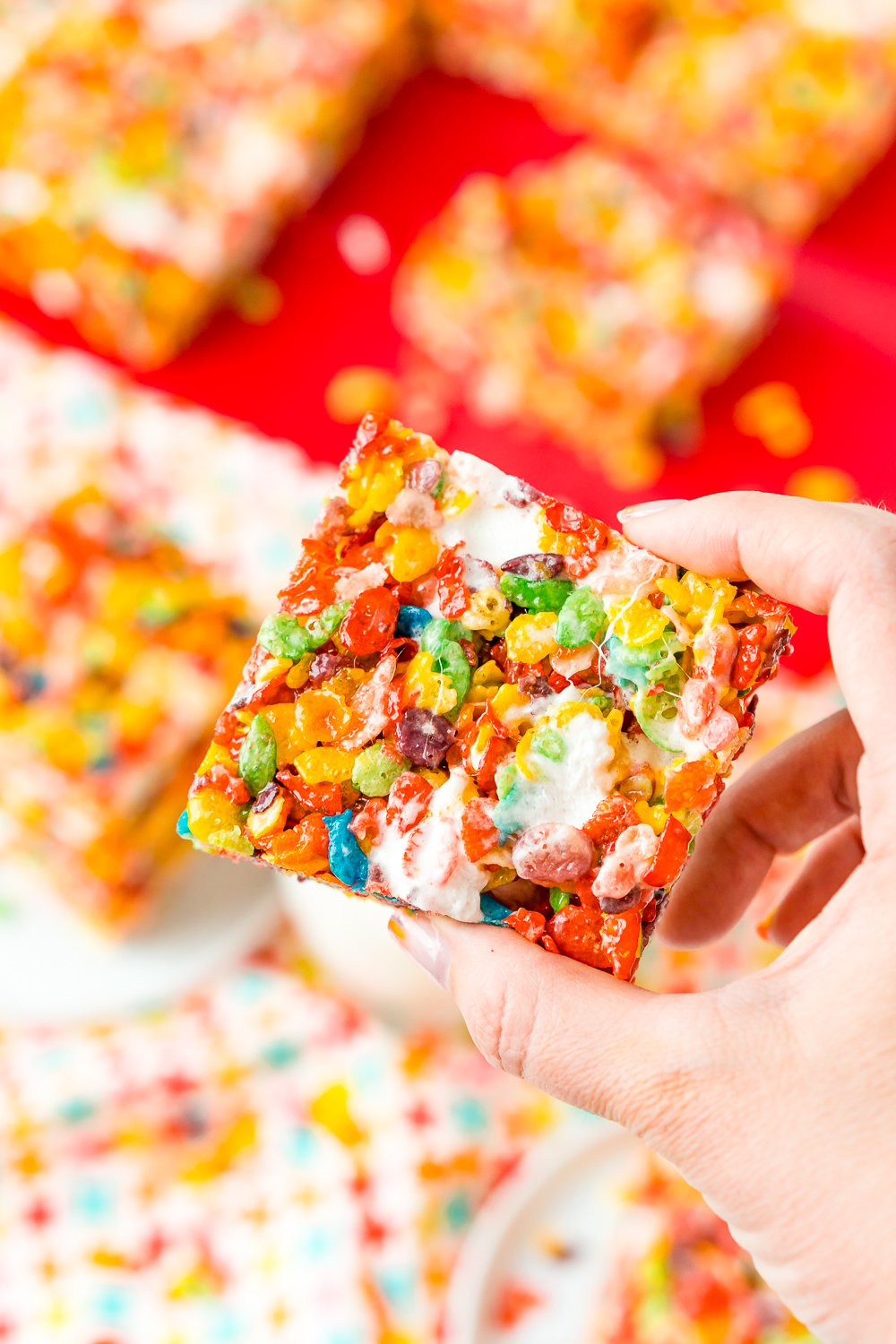 Woman's hand holding a fruity pebbles treat, more treats are in the background on a red cutting board.