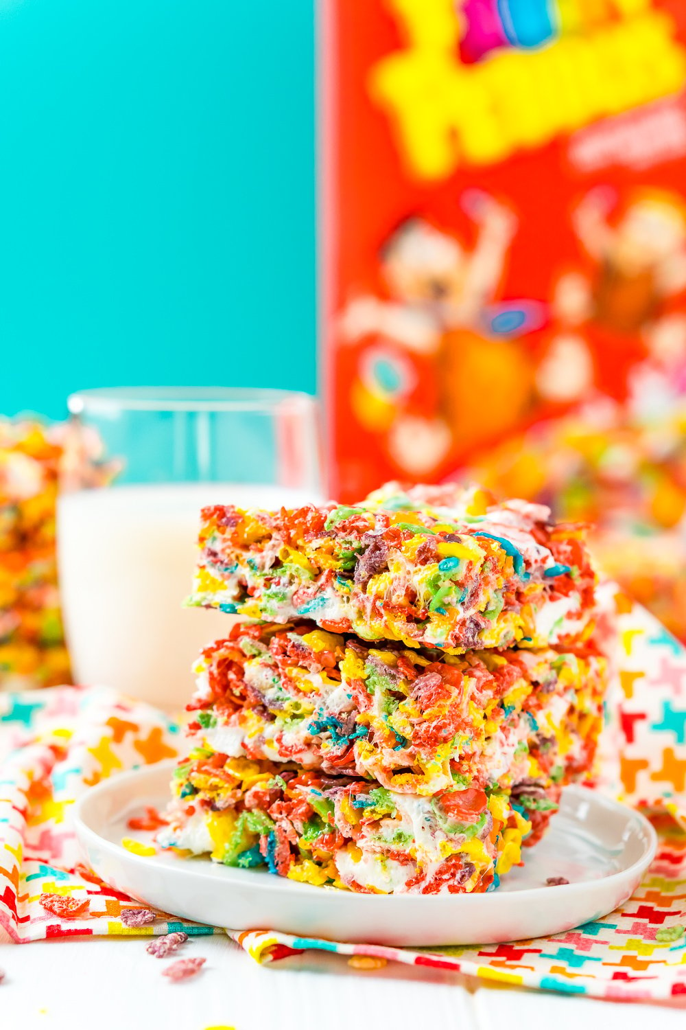 Stack of Fruity Pebbles Treats on a white plate with a glass of milk and a box of fruity pebbles cereal in the background.