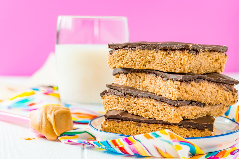 Peanut butter bars stacked on top of each other on a small white plate with spoon of peanut butter next to it and glass of milk in the background.