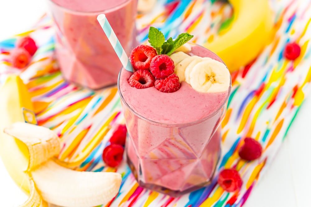 Wide photo of a glass filled with a raspberry banana smoothie with bananas and raspberries on the side.