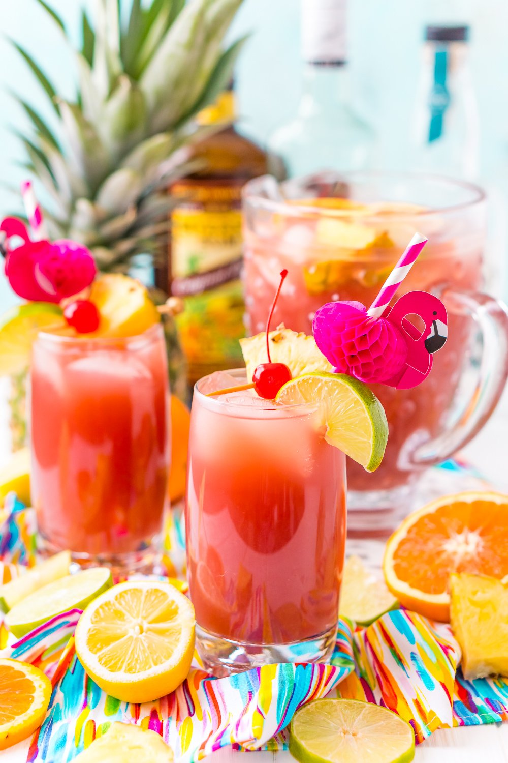 Glasses of rum punch with cherry, pineapple, and lime garnishes. Flamingo paper straws and a pitcher of punch in the background.