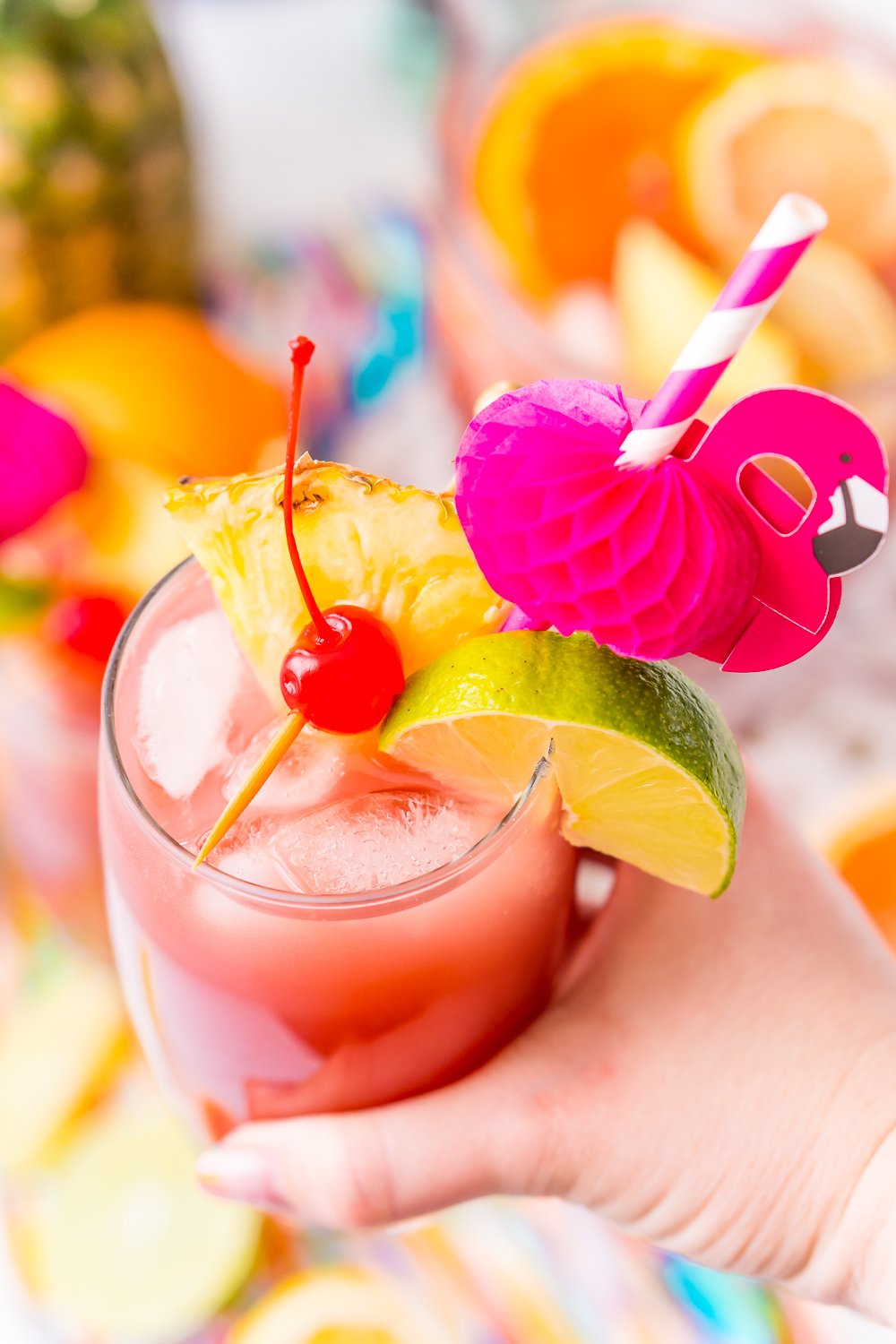 Woman's hand holding a glass of punch that's garnished with a cherry, lime wedge, and pineapple wedge and served with a flamingo paper straw.