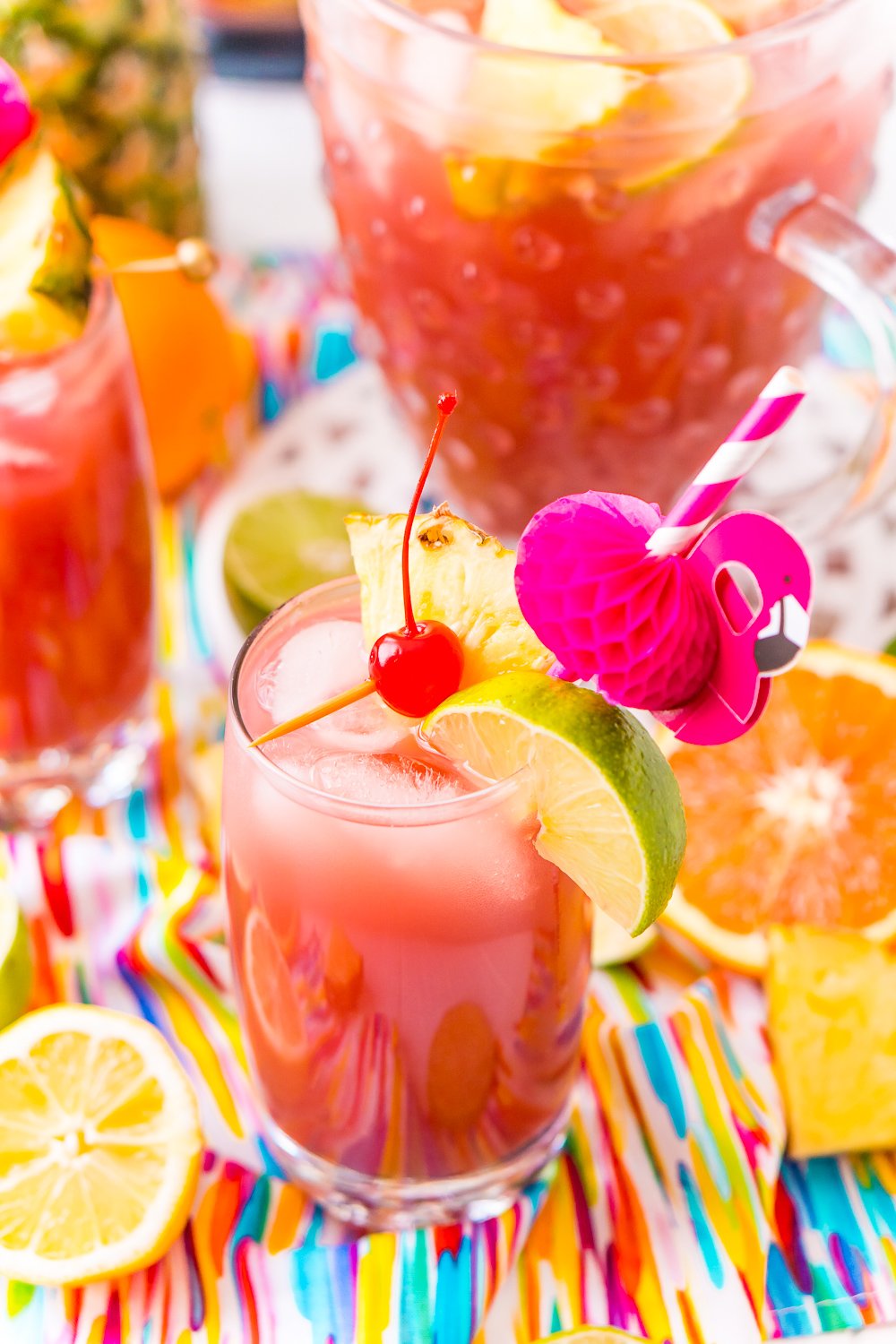 Glass of alcoholic punch garnished with cherry, lime, and pineapple and served with a flamingo straw. Fresh citrus slices scattered around the glass.