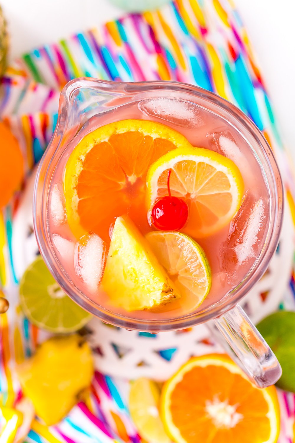 Pitcher of rum punch on a multi-colored napkin with fruit slices on the counter.