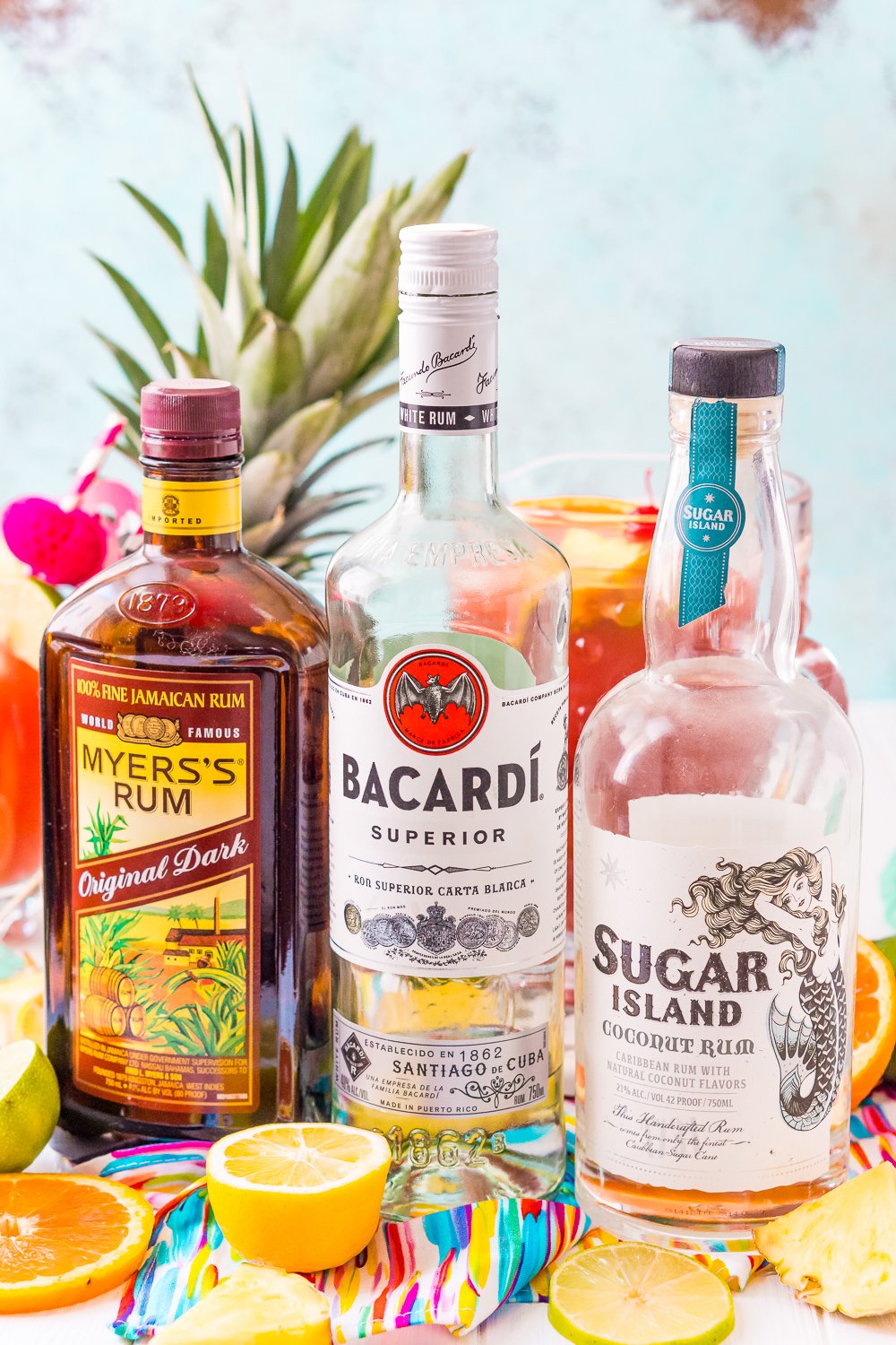 Bottles of Myer's Rum, Bacardi, and Sugar Island Rum lined up with a pitcher of rum punch and a pineapple in the background.