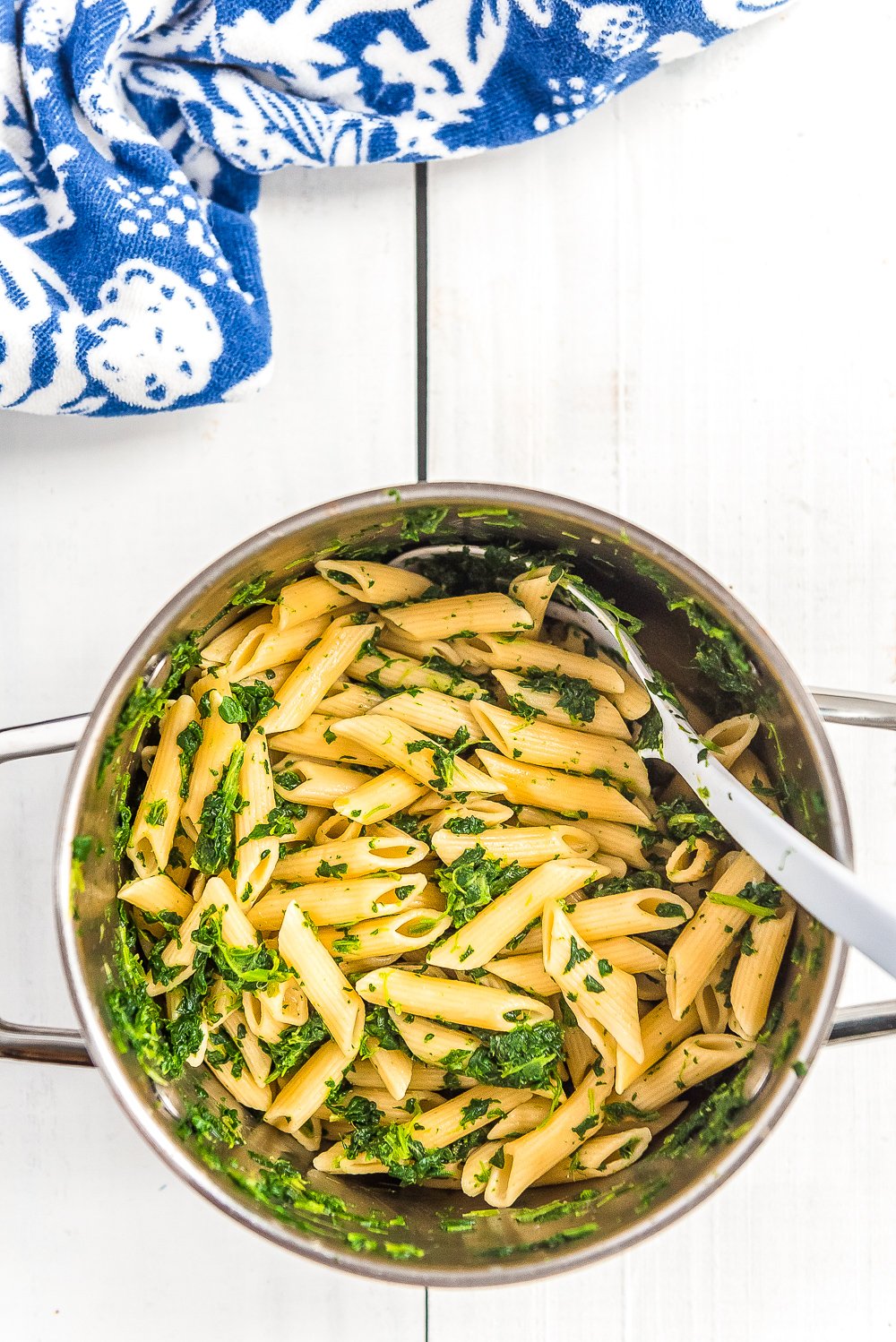 Cooked pasta in a pot with spinach on white wood table with blue and white napkin.