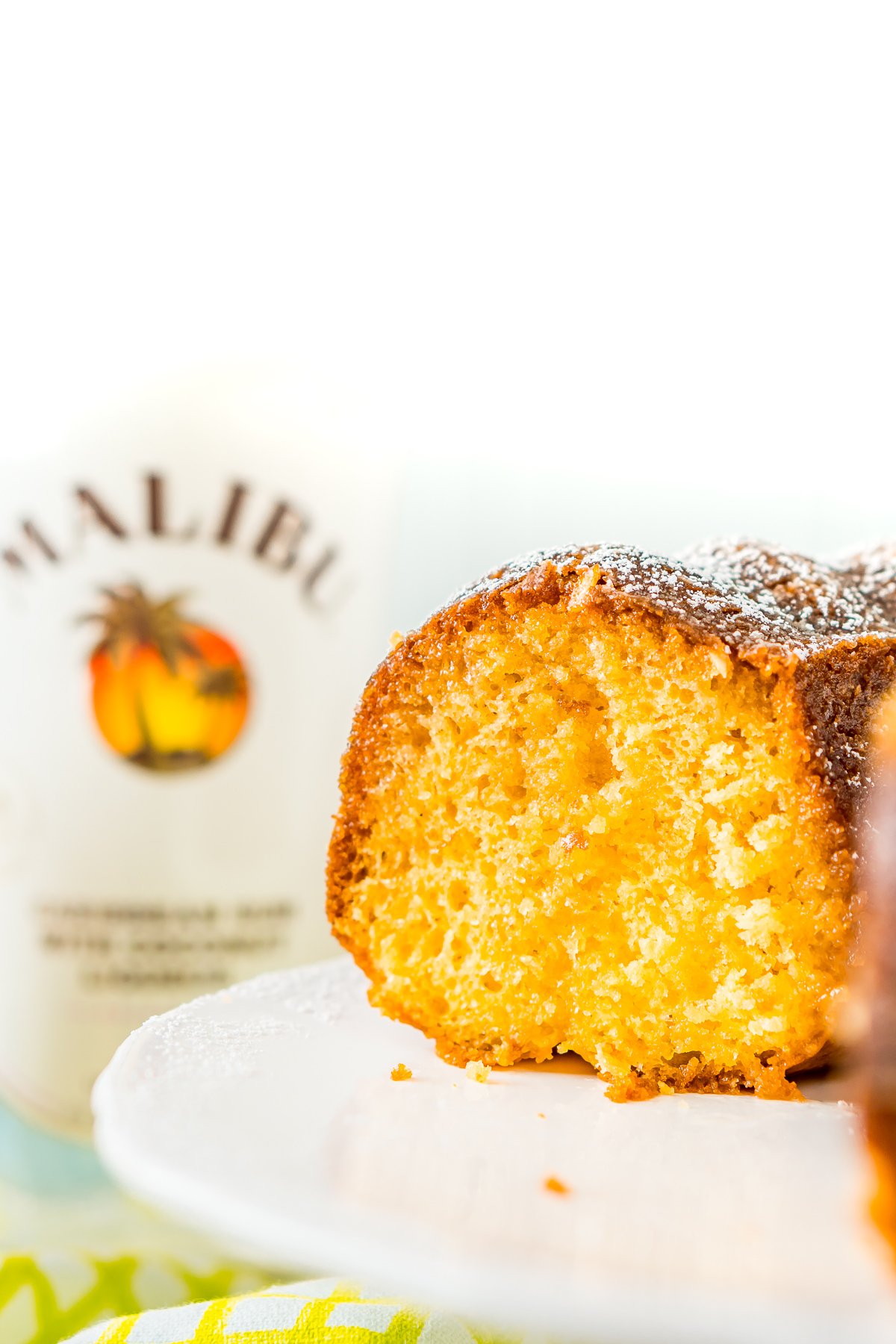 Rum cake with a slice taken out of it on a cake stand with a bottle of coconut rum in the background.