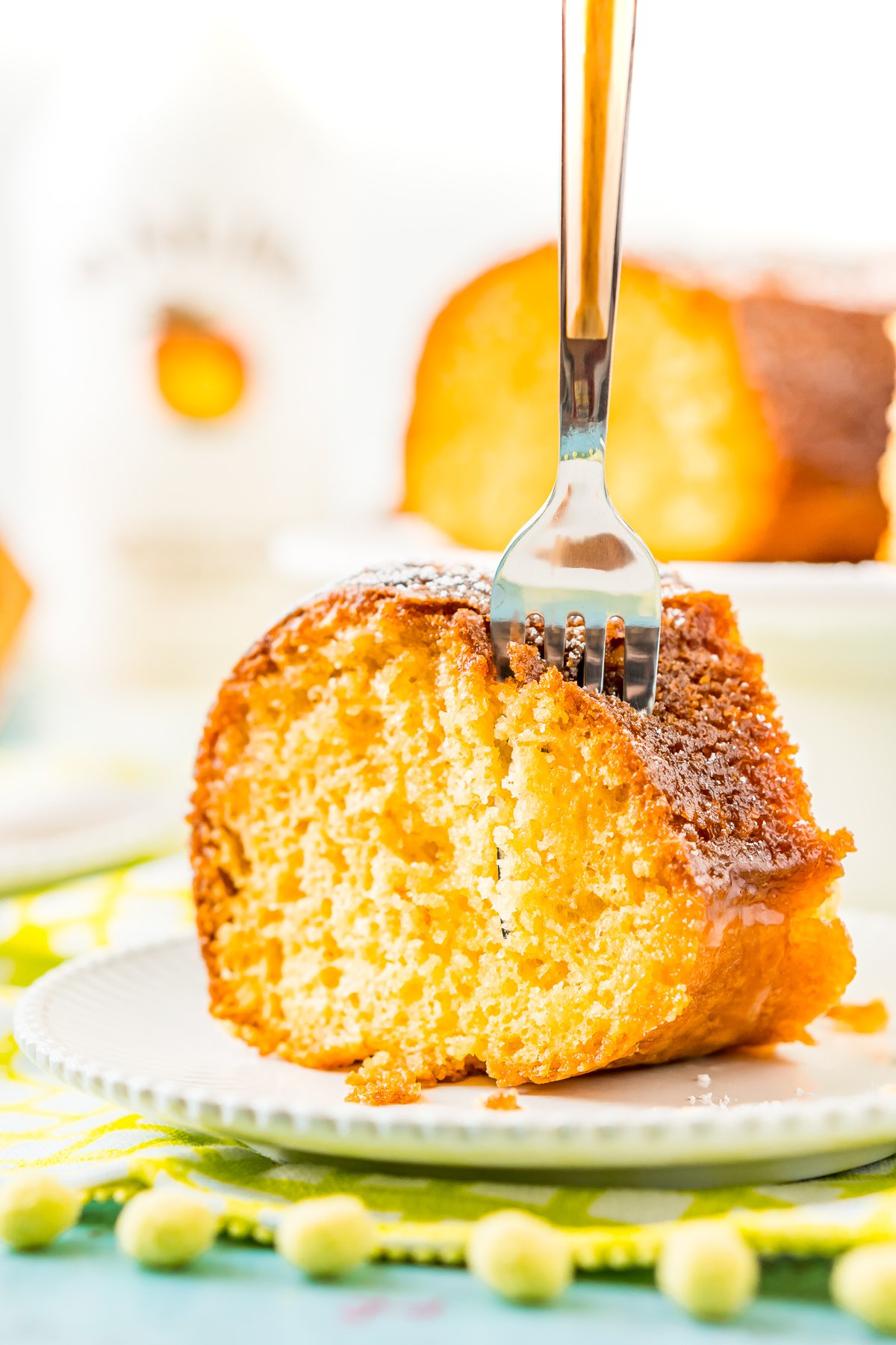 Fork taking a bite out of a slice of coconut rum cake on a white plate with a bottle of rum and more cake in the background.