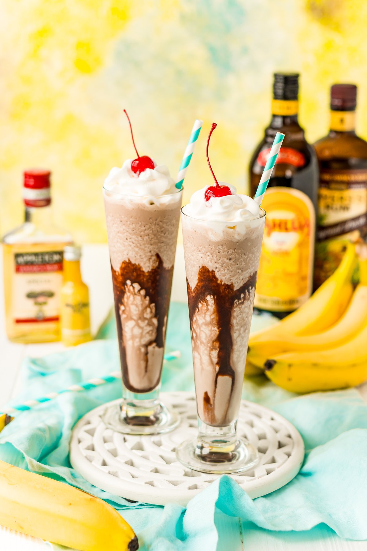 Two dirty banana drinks on a white wooden board with bottles of rum and bananas in the background.