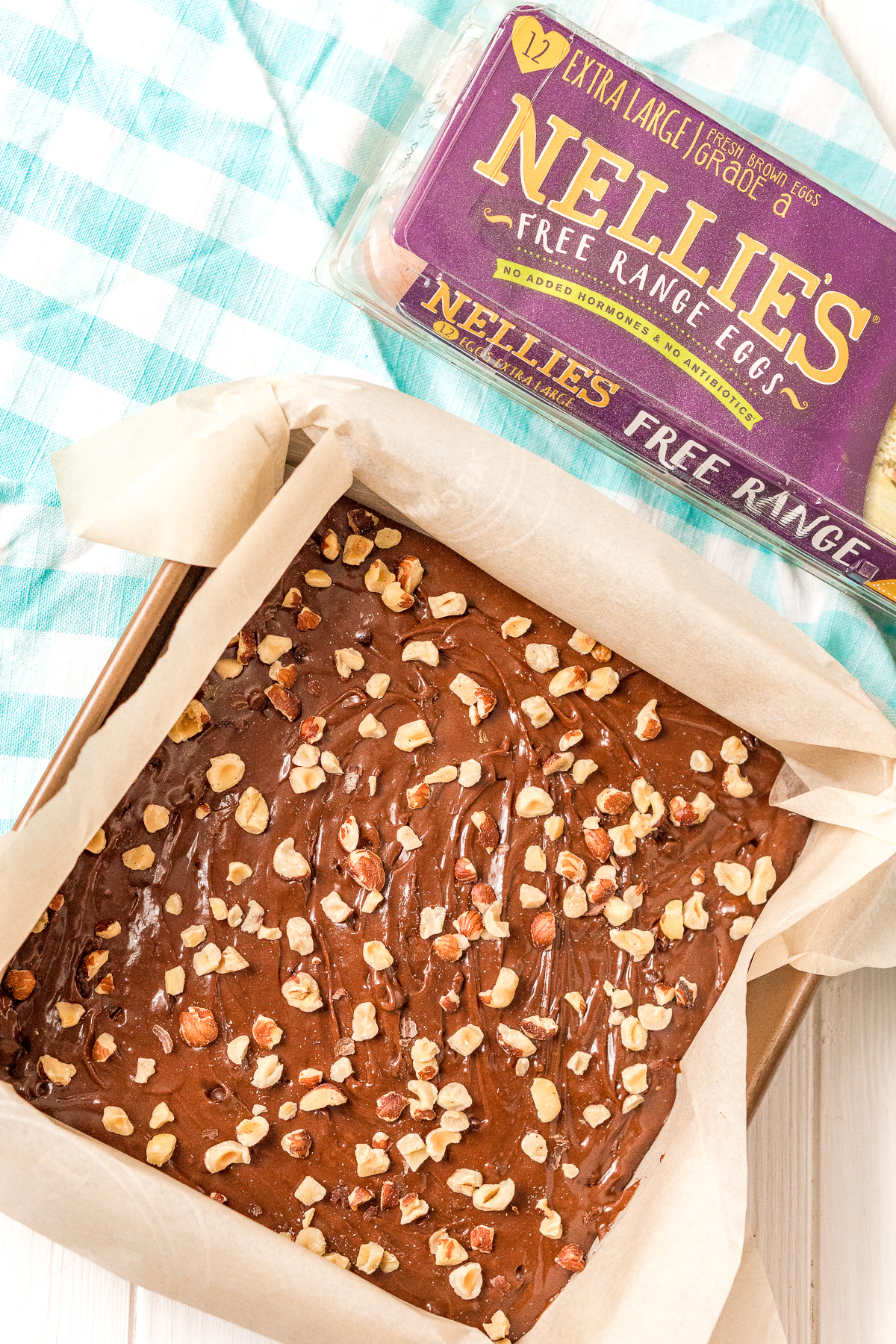 Uncooked brownie batter in a square baking dish topped with chopped hazelnuts next to a carton of eggs.