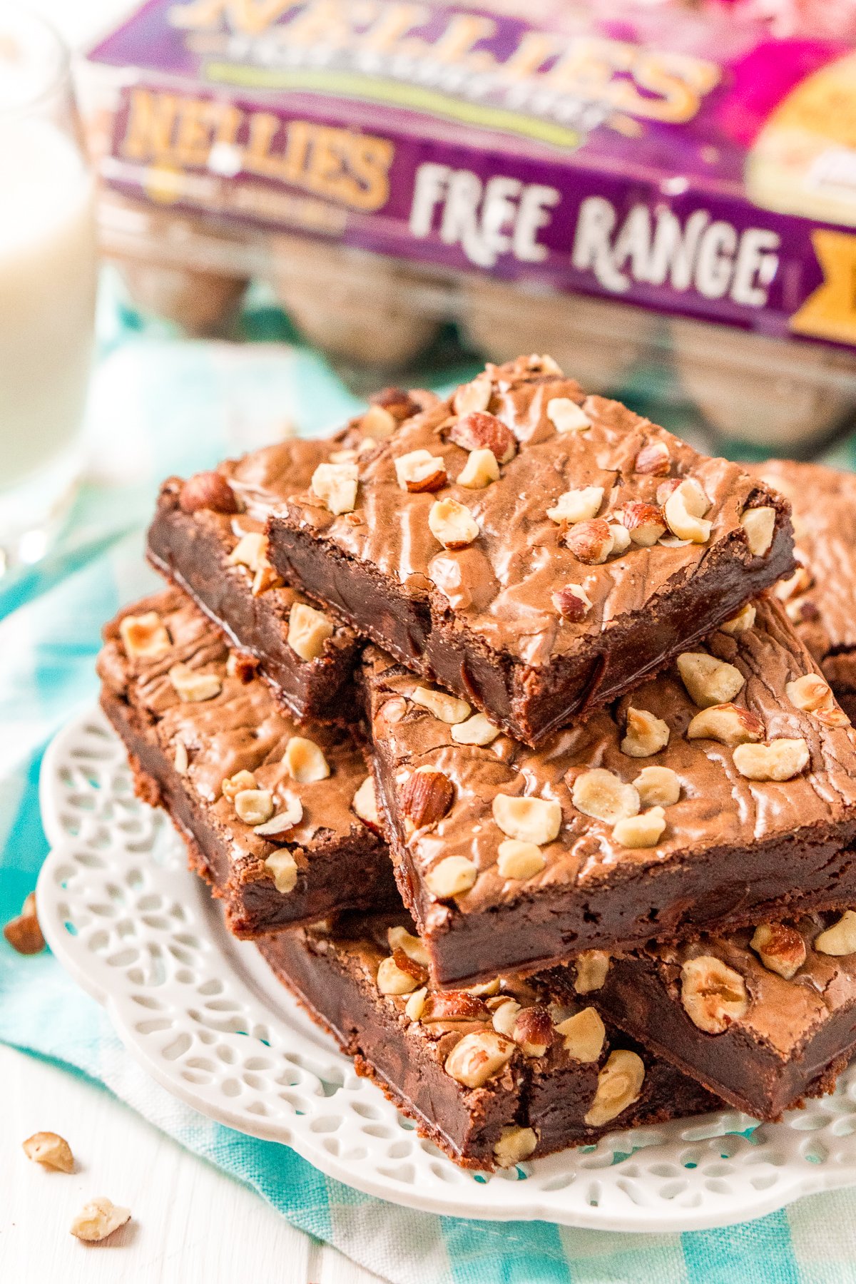 Brownies with hazelnuts stacked on a white plate with a carton of eggs in the background.