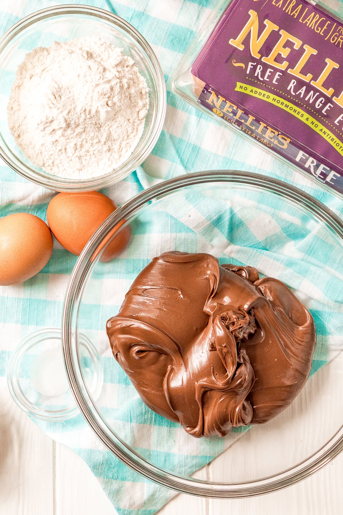 Nutella in a glass bowl with carton of eggs, loose eggs, small bowl of flour and salt on a white and blue napkin.