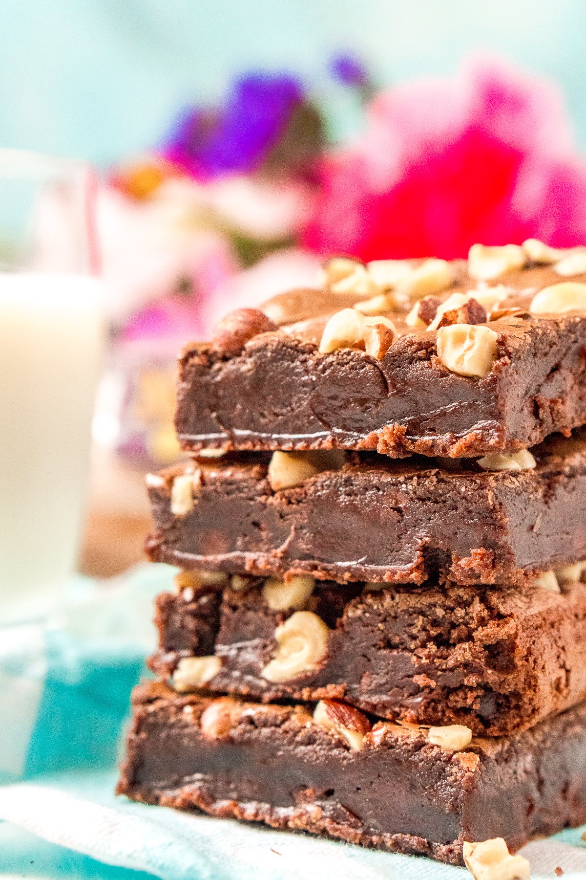 Stack of four brownies with hazelnuts. Glass of milk and bouquet of flowers in the background.