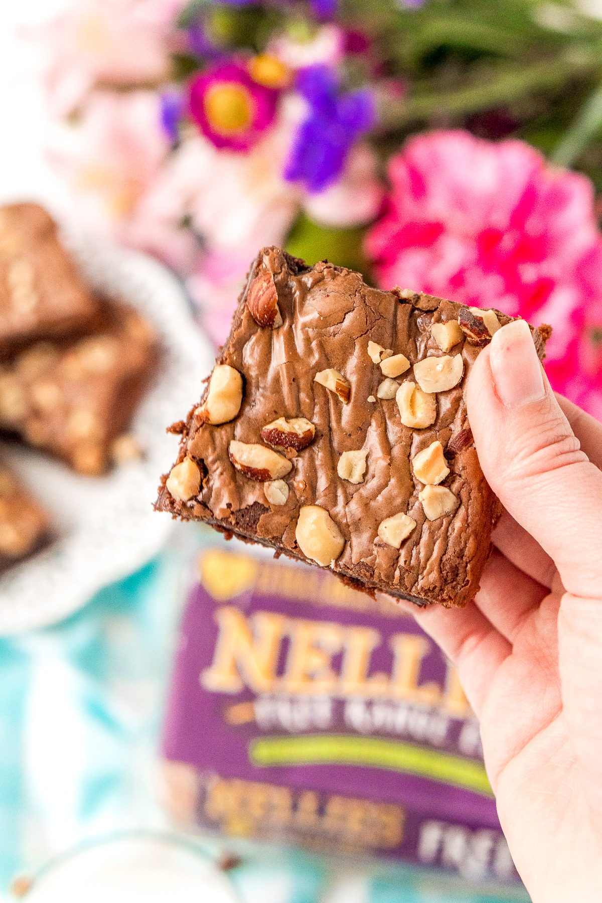 Woman's hand holding a hazelnut brownie. More brownies and flowers in the background.