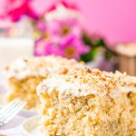 Slice of cake on a small white dessert plate with a pink background and flowers in the background.