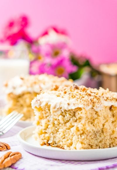 Slice of cake on a small white dessert plate with a pink background and flowers in the background.