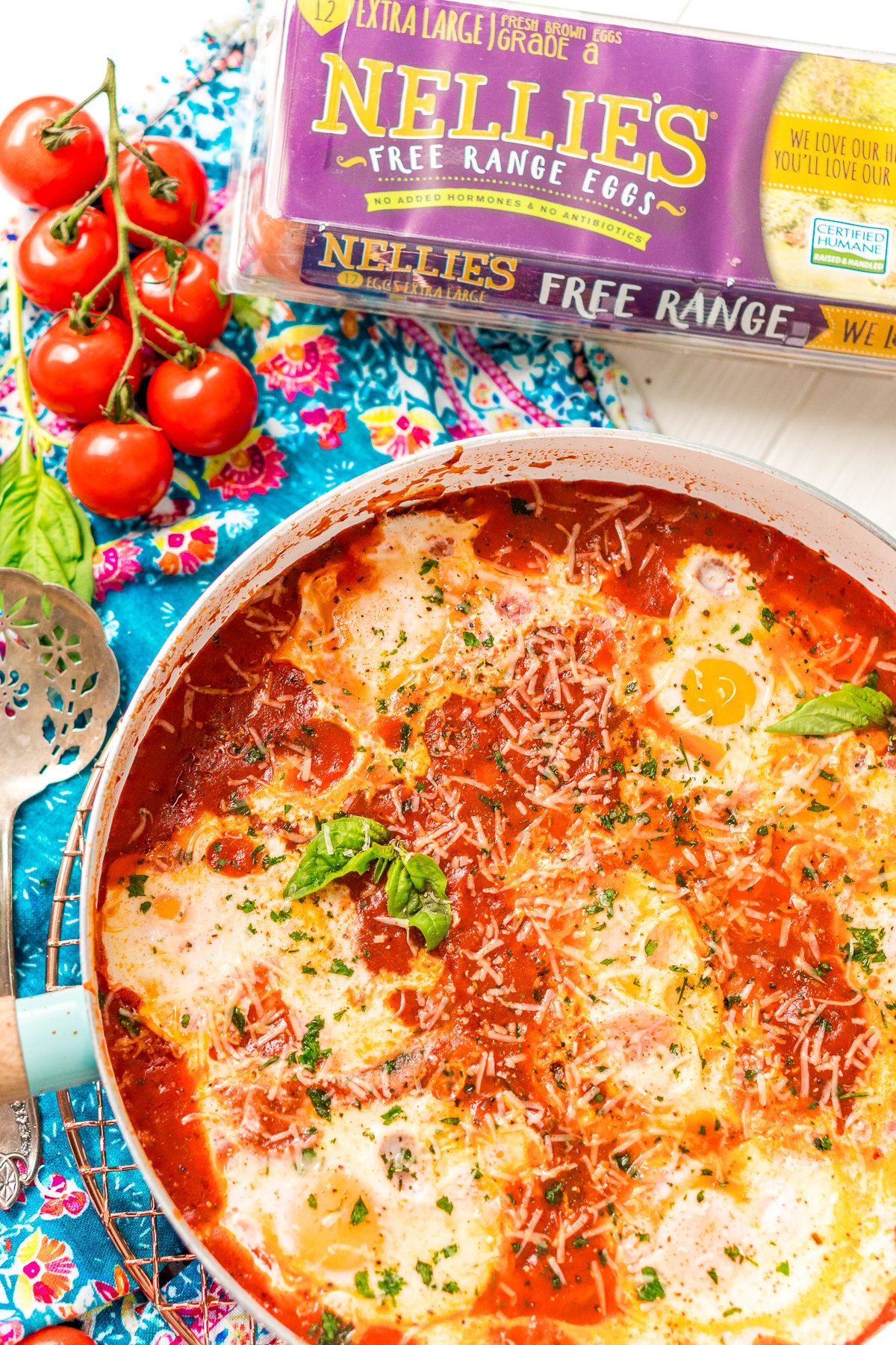 Eggs In Purgatory in a white and blue skillet with a blue napkin. Tomatoes and a carton of eggs are to the side of the pan.