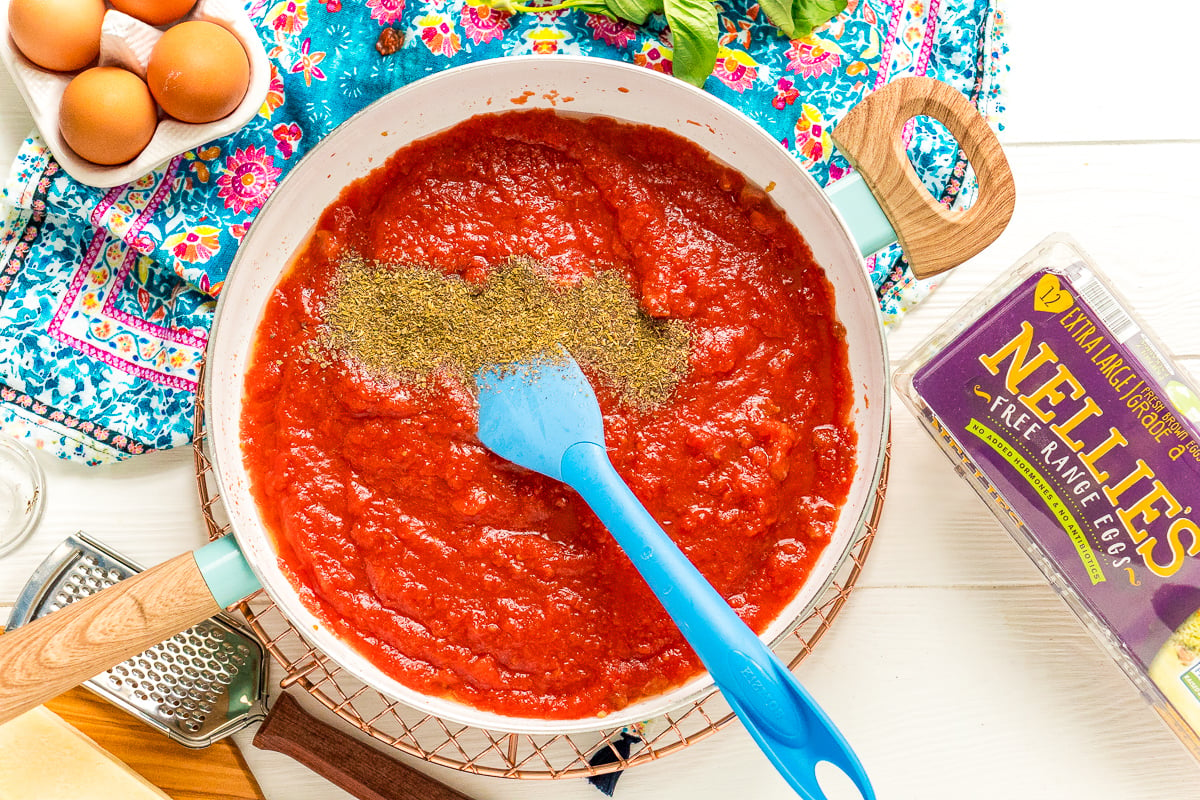 Italian Seasoning being added to a fry pan with tomato sauce.