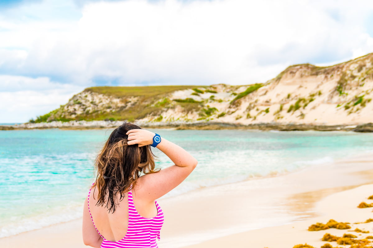 Woman standing back to on a beach in a pink swimsuit with hand in her hair.