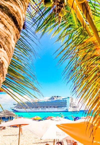 Photo of a Princess Cruise Ship in port. Palm trees bordering the photo in the foreground. Beach umbrellas.