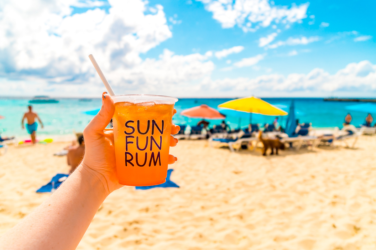 Woman's hand holding a cup of rum punch on a beach in front of beach umbrellas.