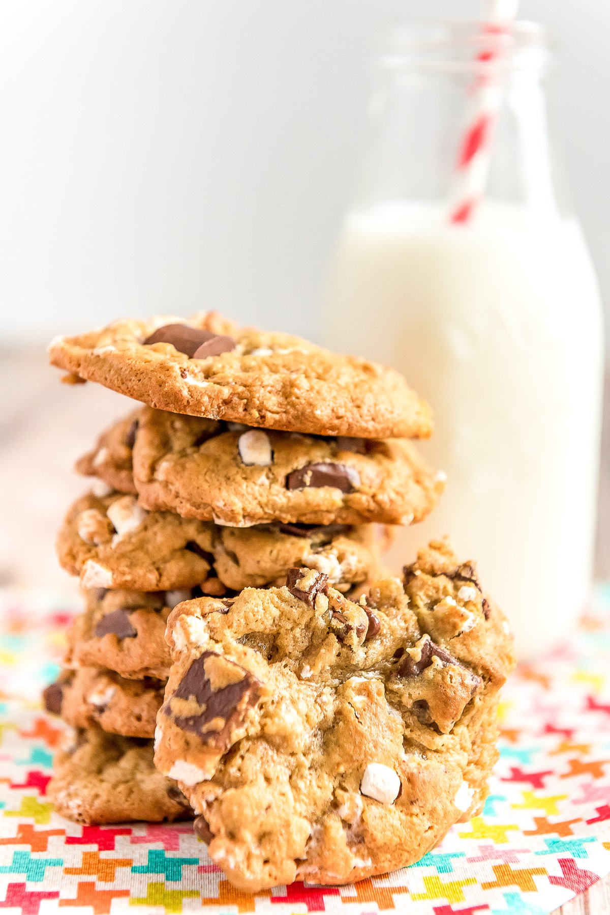 Stack of cookies with marshmallows and chocolate chunks in front of a glass of milk. One cookie is leaning against the others.