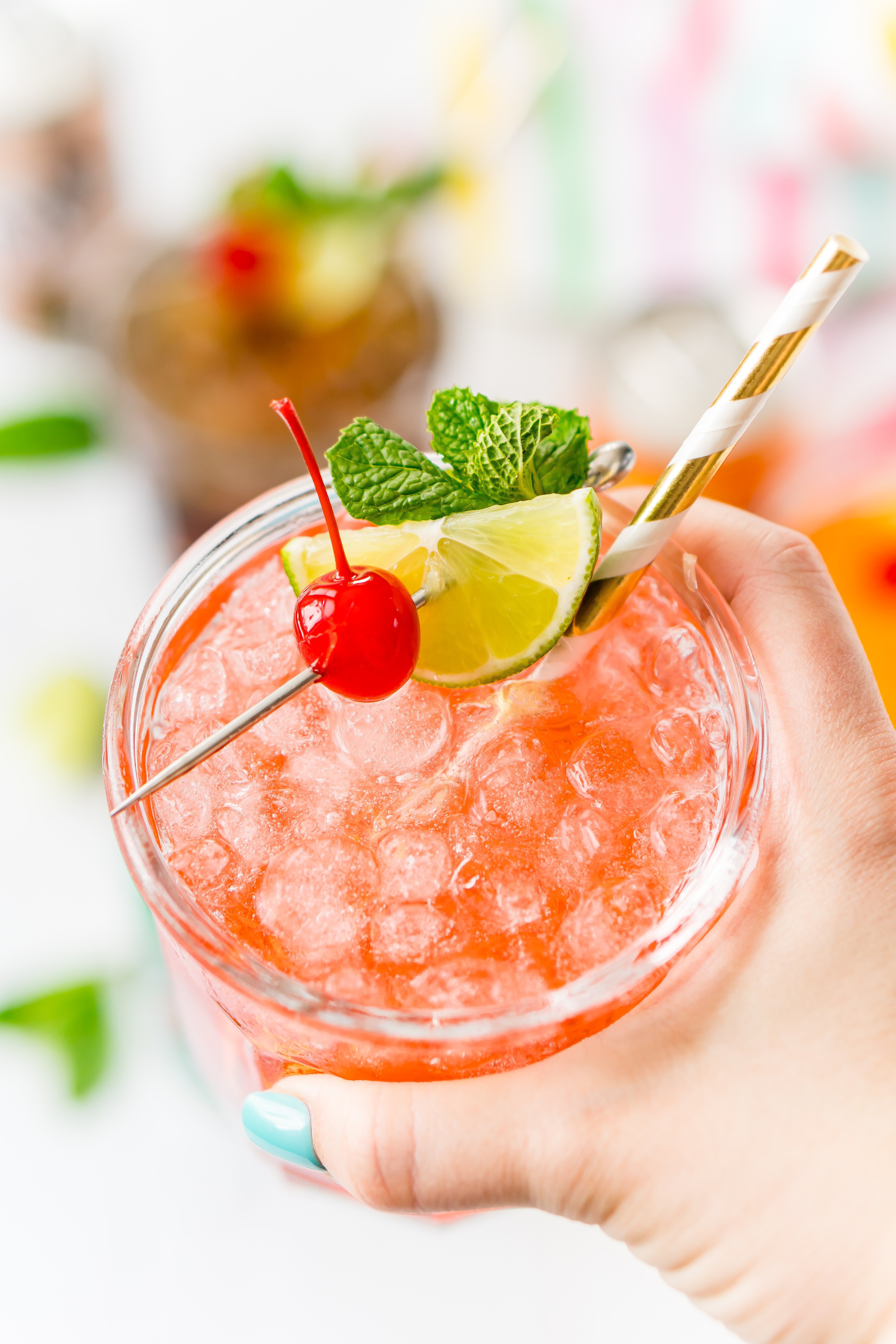 Woman's hand holding up a drink garnished with cherry, mint, and lime.